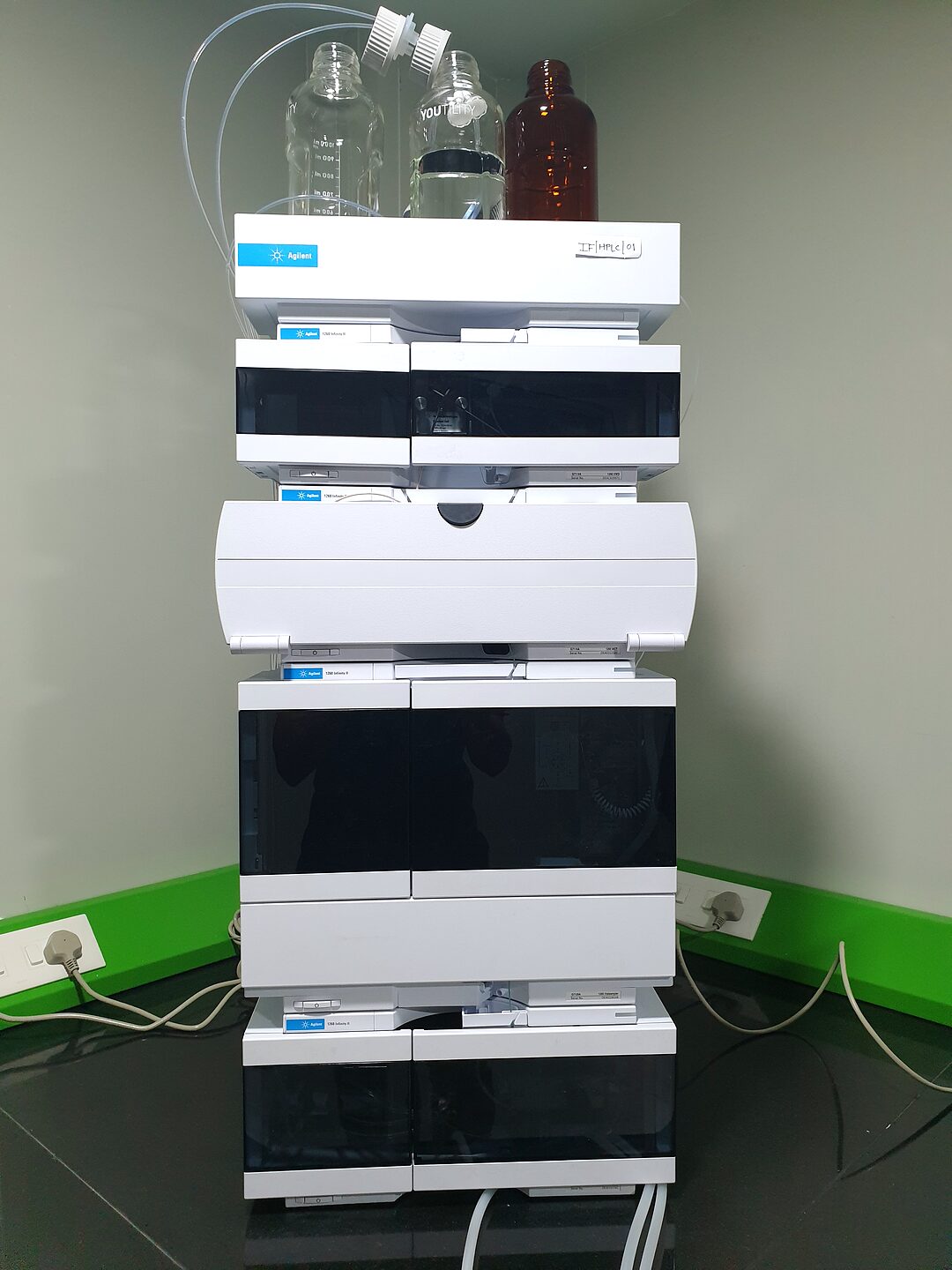 An Agilent HPLC stack with solvent bottles on top, tagged IF/HPLC/01, standing against a green laboratory wall.